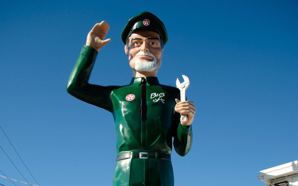 Un géant en fibre de verre en uniforme vert, employé de station-service, qui salue en tenant une clé à molette
