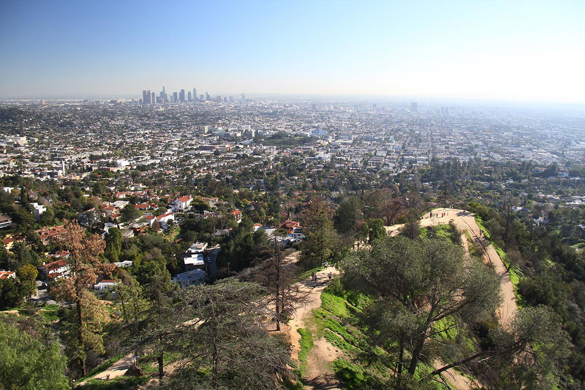Los Ángeles extendiéndose hasta el horizonte desde las colinas de Griffith Park, con las torres del centro a lo lejos y excursionistas en un sendero más abajo