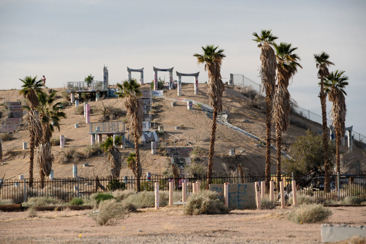 El parque acuático abandonado de Lake Dolores: torres, escaleras y pilares cubiertos de grafitis en una colina del desierto tras una valla y palmeras desgreñadas