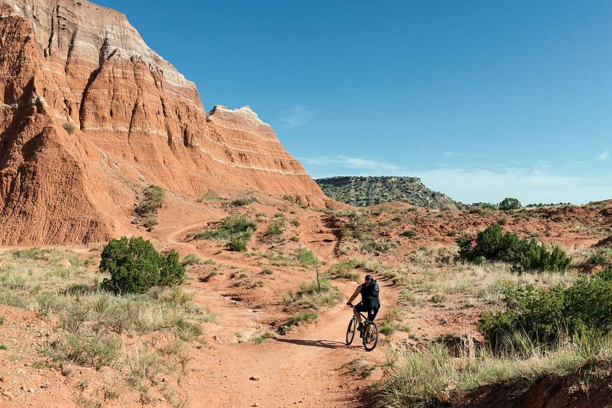 Un ciclista en un sendero de tierra roja bajo una formación rocosa de rayas rojas y blancas en Palo Duro Canyon