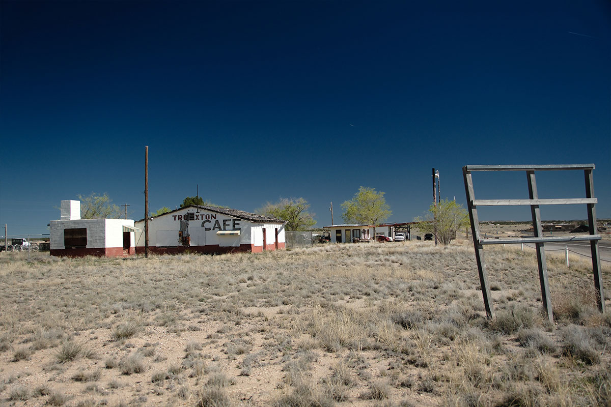 El Truxton Café cerrado, un edificio blanco con adornos rojos y letras pintadas, junto a un marco de letrero vacío sobre pastizal seco